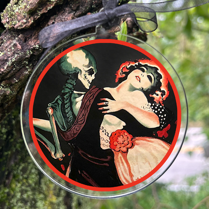 A round glass ornament featuring  a skeleton and a woman, hanging from a black ribbon on a mossy tree branch in a forest.