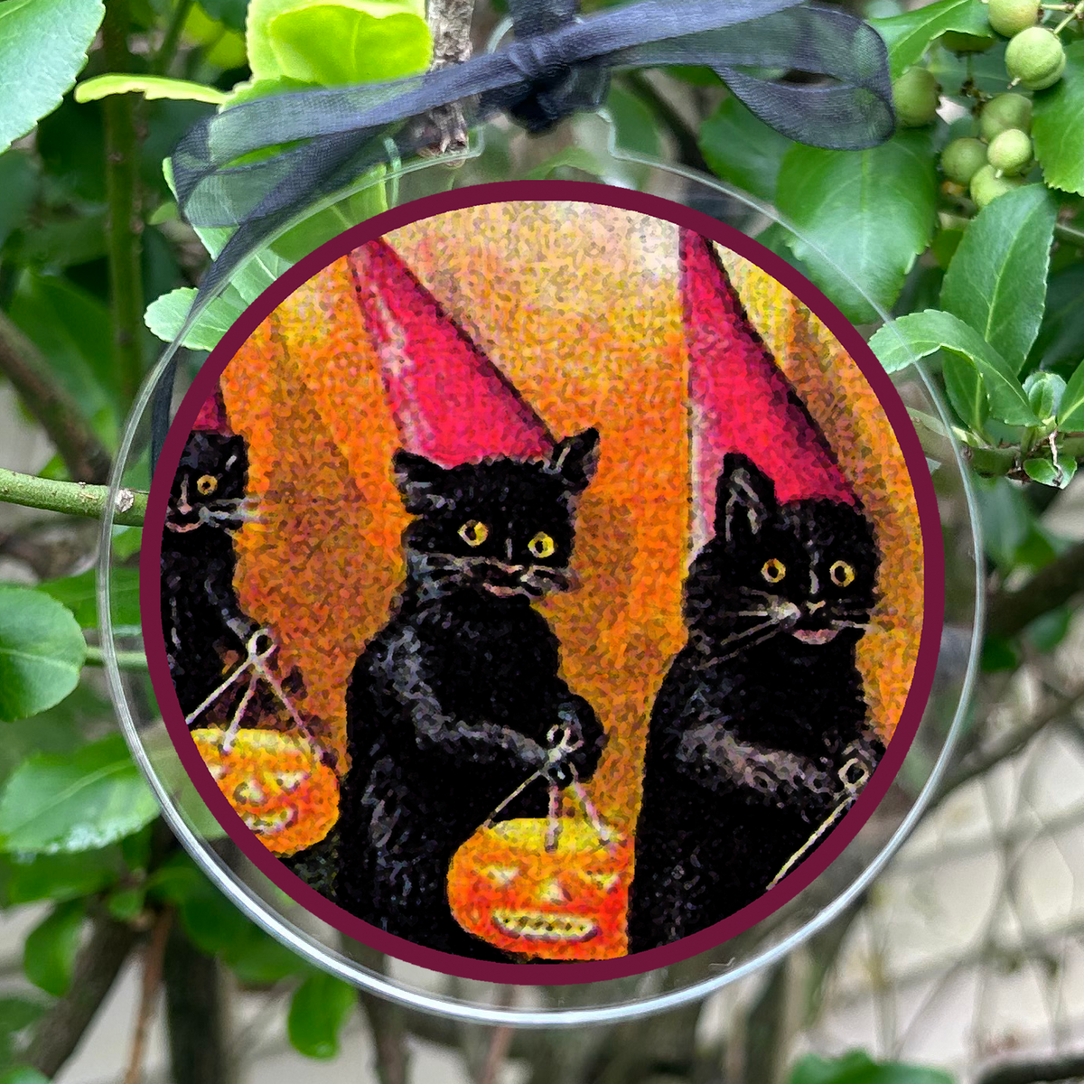 A round clear acrylic hanging ornament featuring three black cats in witch hats with jack-o'-lanterns, suspended by a black ribbon among green leaves and small green berries.