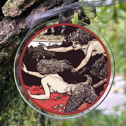 A round glass ornament with a black ribbon hanging from it, featuring an antique-style illustration of two figures in a red-bordered frame, photographed outdoors against a blurred forest background.