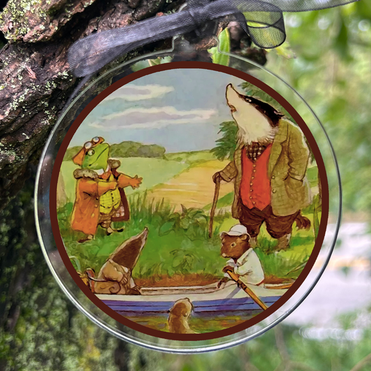A round hanging ornament with a black ribbon, featuring an illustrated scene of anthropomorphic animals by a river, photographed hanging from a tree branch in a forest.