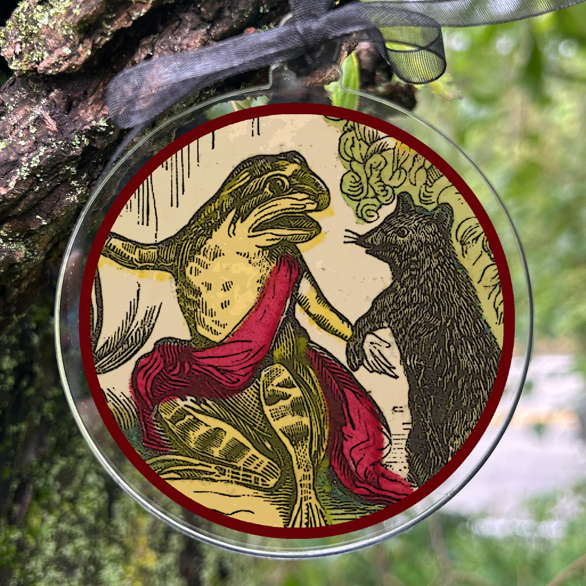 A round clear glass hanging ornament with a printed vintage-style illustration children’s book The Little Frog and Pretty Mouse by American artist John B. Keller. suspended by a sheer gray ribbon from a tree branch outdoors.