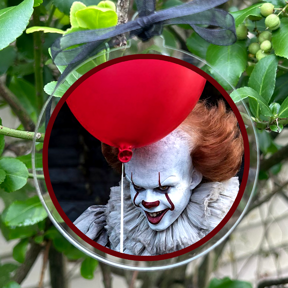 A round clear glass hanging ornament featuring Pennywise the Clown holiday a red balloon. The ornament is suspended by a black ribbon from a leafy green tree branch.