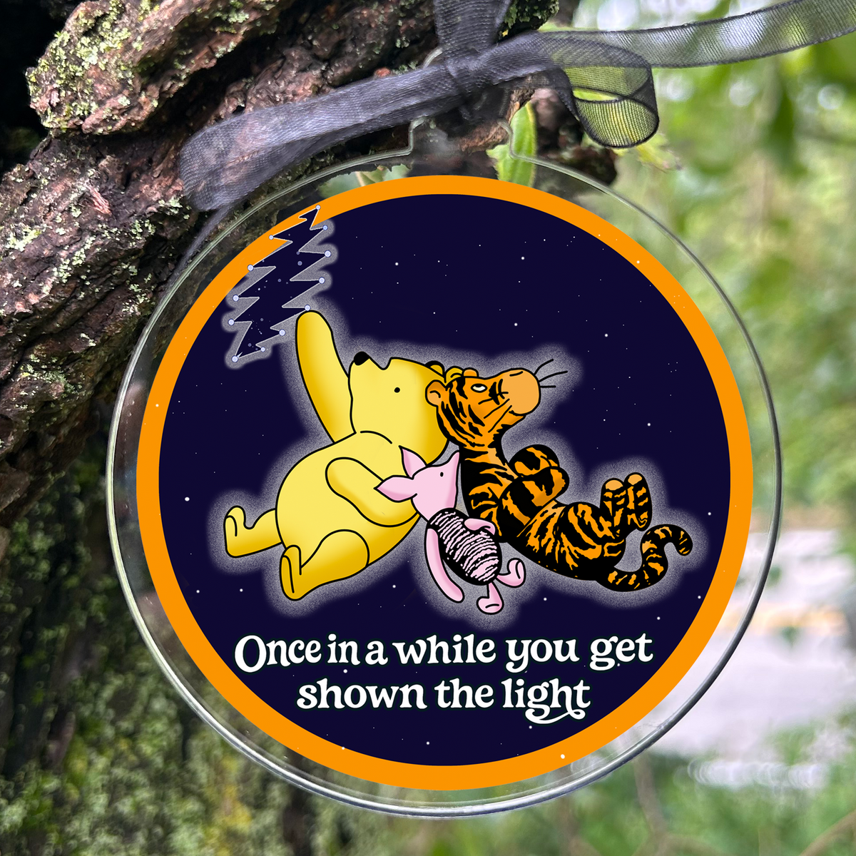 A round glass ornament hanging from a black ribbon on a tree trunk outdoors. The ornament features a cartoon illustration of Winnie the Pooh and Tigger on a dark blue background with an orange border, and the text 'Once in a while you get shown the light' at the bottom.
