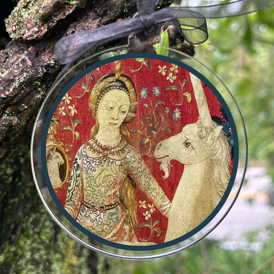 A round hanging ornament with a clear border and black ribbon, featuring a medieval-style illustration of a woman in gold clothing beside a white unicorn on a red background, photographed hanging from a tree trunk outdoors.