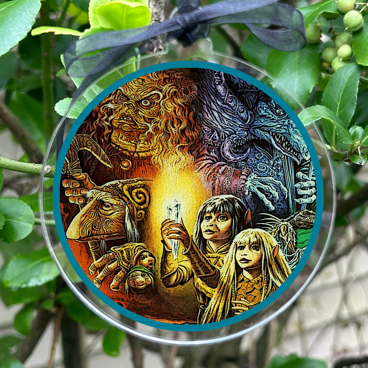 A circular glass ornament featuring an illustrated design of Gelflings Kira & Jen , suspended by a sheer black ribbon among green leaves and small berries.