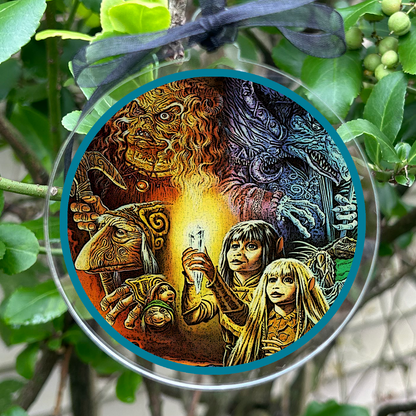 A circular glass ornament featuring an illustrated design of Gelflings Kira & Jen , suspended by a sheer black ribbon among green leaves and small berries.
