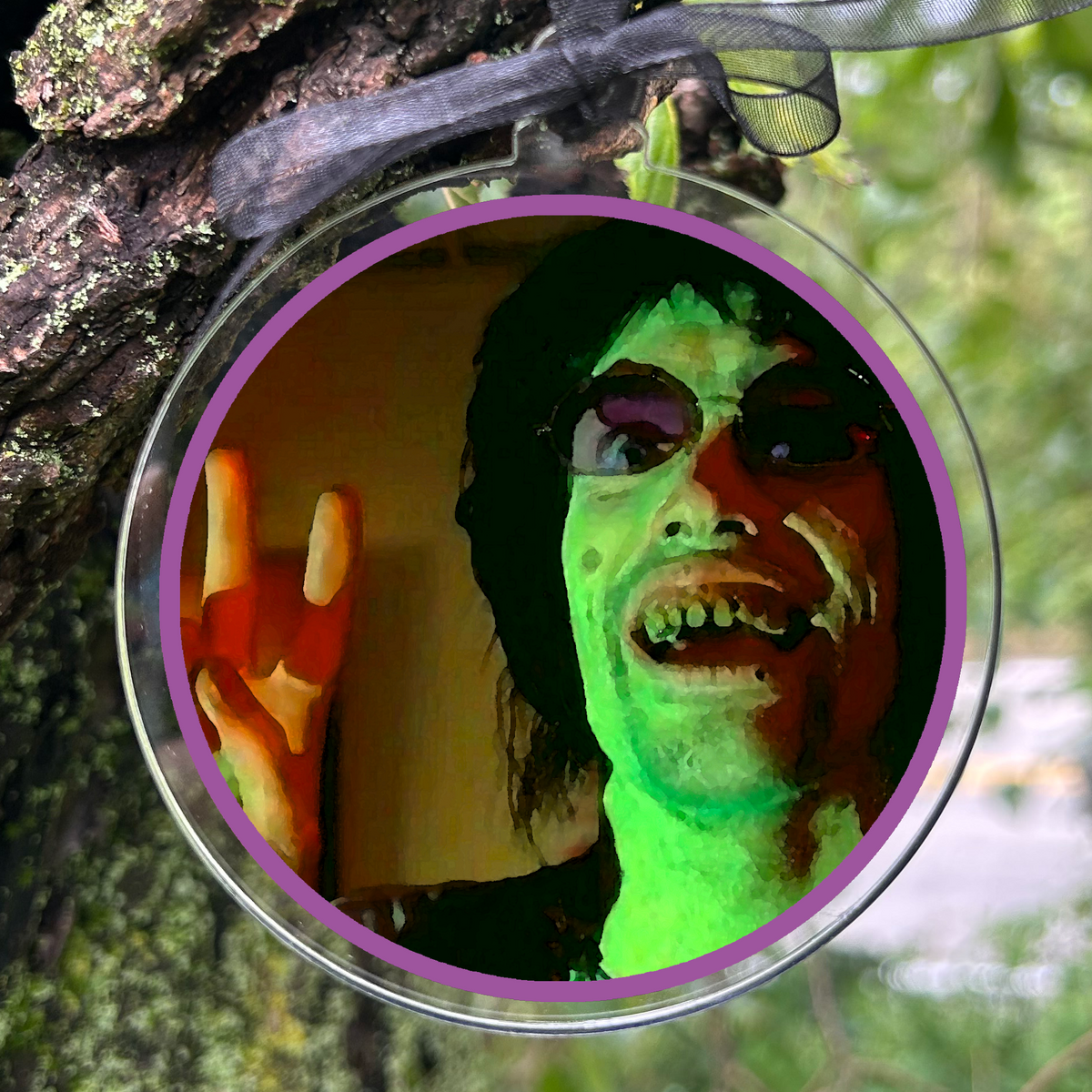 A round clear hanging ornament with a printed photo of a person with neon green skin and a black ribbon, photographed hanging from a tree branch outdoors.
