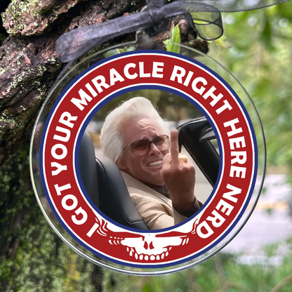 A round clear plastic ornament with a printed photo of an older man in a car and bold red and white text reading 'I GOT YOUR MIRACLE RIGHT HERE NERD', hanging from a black ribbon on a tree branch outdoors.