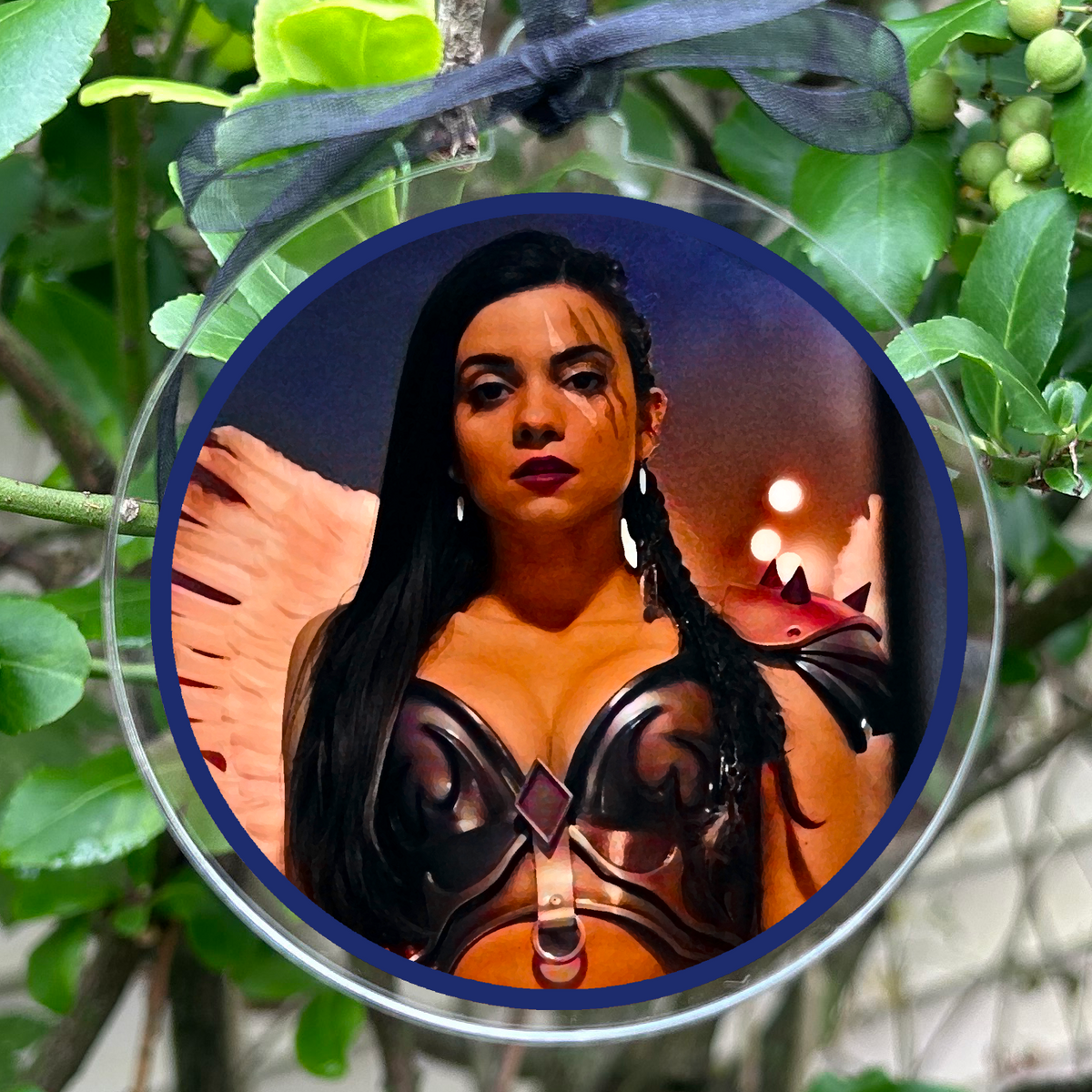 A round hanging ornament with a clear circular frame and a black ribbon loop, featuring a printed portrait of a woman in a black and orange outfit. The ornament is photographed hanging among green leaves and small berries outdoors.