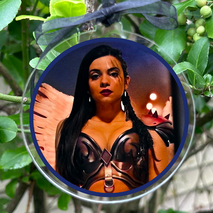 A round hanging ornament with a clear circular frame and a black ribbon loop, featuring a printed portrait of a woman in a black and orange outfit. The ornament is photographed hanging among green leaves and small berries outdoors.