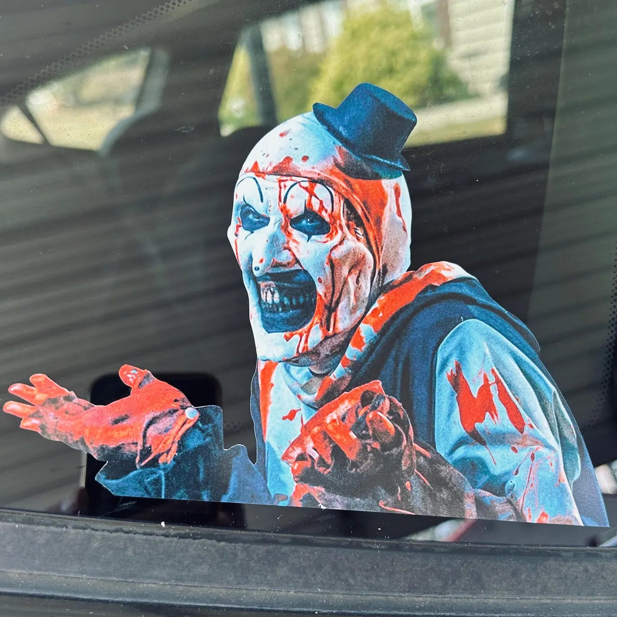 A die-cut vinyl decal of a horror Art the  clown with a bloody face, blue top hat, and blue-and-red outfit, shown applied to a car window.
